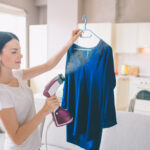 Woman,Is,Steaming,Blue,Shirt,In,Room.,She,Holds,Small